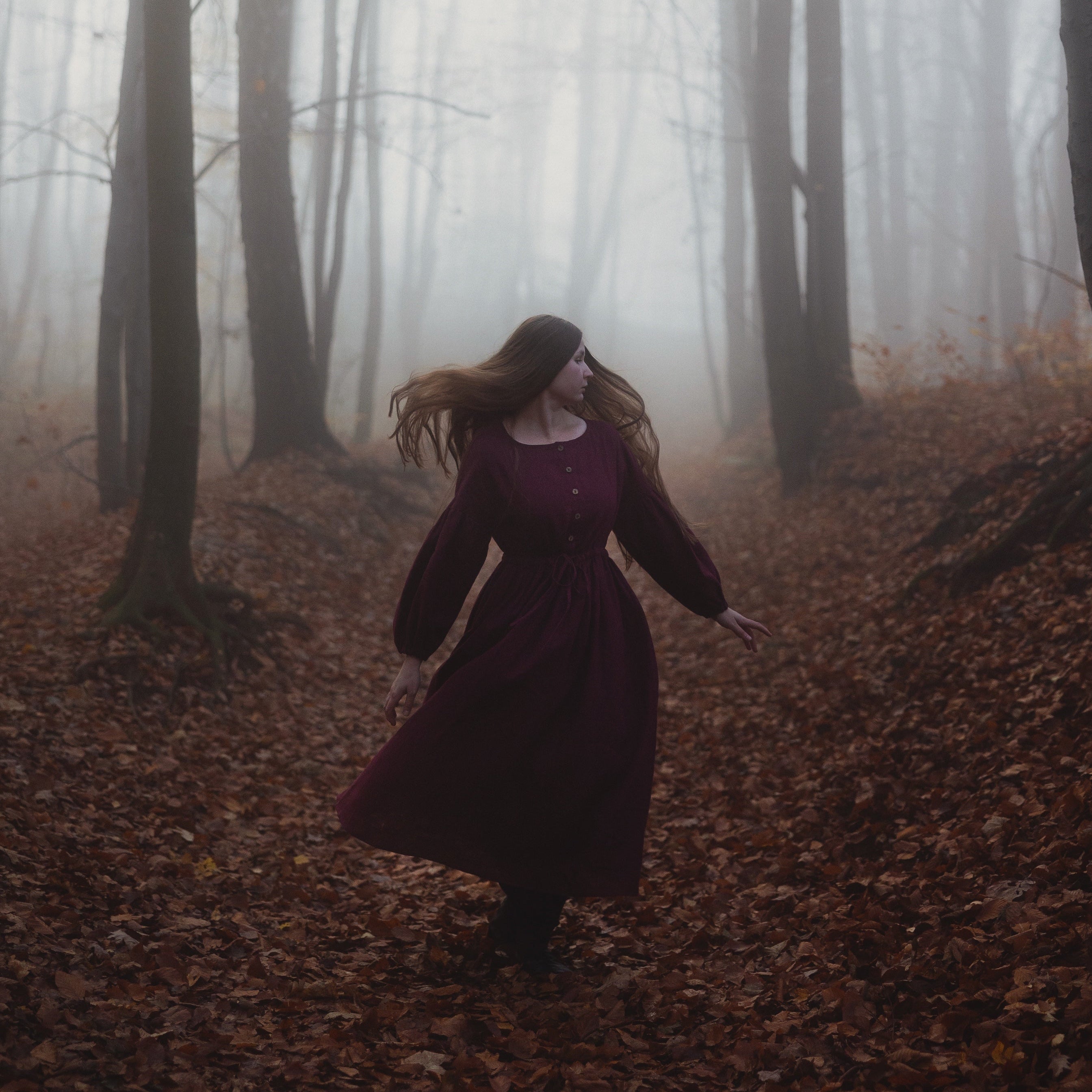 A woman in a long dark plum dress walking through a misty forest path covered with brown fallen leaves.